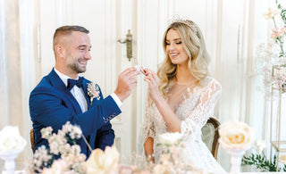 Nevěsta a ženich sedí u stolu a s úsměvem společně ochutnávají makronky, v pozadí svatební výzdoba.