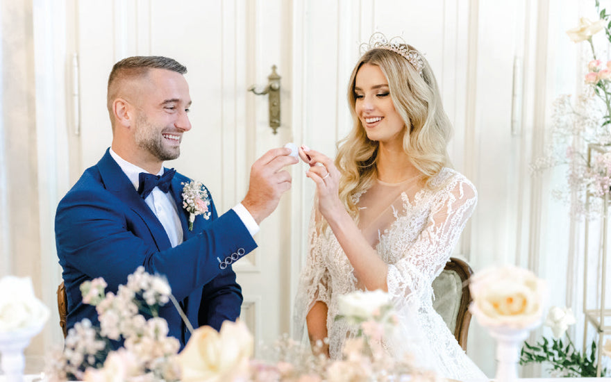 Nevěsta a ženich sedí u stolu a s úsměvem společně ochutnávají makronky, v pozadí svatební výzdoba.